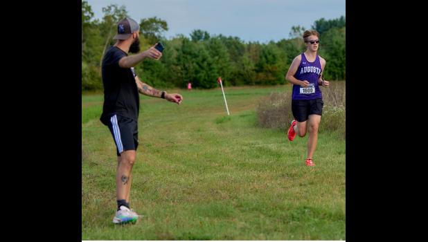 Augusta's Thomas Truog runs on the backstretch of the AHS course in the Augusta Invitational meet encouraged by his coach. Photo by James Truog